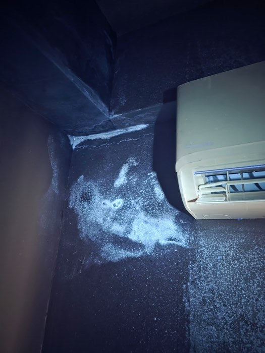 The wall beside the small bedroom air-conditioner in the Bandar Sri Damansara flat under ultraviolet light, a heavy fluorescent halo and condensate streaks tracking down